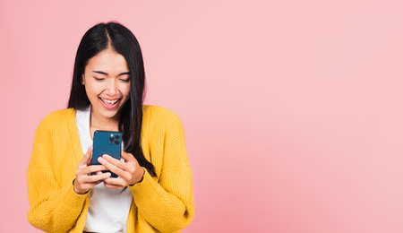 Happy Asian portrait beautiful cute young woman teen smiling excited using smart mobile phone studio shot isolated on pink background, Thai female surprised making winner gesture on smartphoneの写真素材