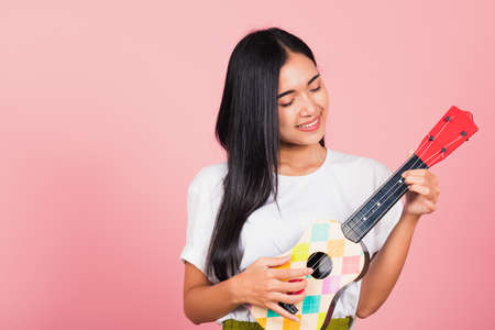 Portrait of happy Asian beautiful young woman teen confident smiling face hold acoustic Ukulele guitar, female playing Hawaiian small guitar, studio shot isolated on pink background, with copy spaceの写真素材