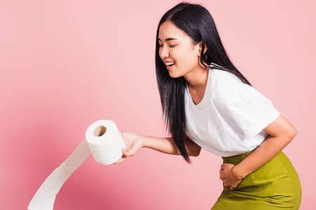 Portrait of Asian beautiful young woman diarrhea constipation holding stomachache and tissue toilet paper roll on hand she crotch lower abdomen want to WC, studio shot isolated on pink backgroundの写真素材