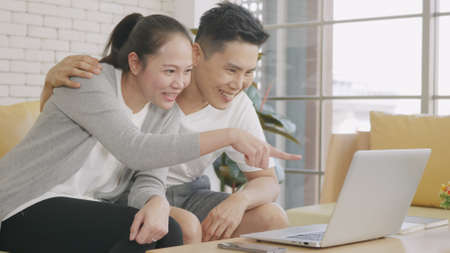 Happy Asian family couple husband and wife laughing sitting on sofa using laptop computer webcam technology talking making online social distance video call to their children at home.の写真素材