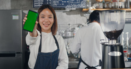 Asian happy beautiful young woman standing behind the bar counter in cafe smiling showing smartphone blank green screen looking camera, small business owner coffee shopの写真素材