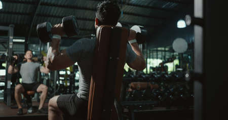 Black view of muscular man guy lifting two dumbbell during workout while sitting on bench, Bodybuilder working out with dumbbell heavy weights at fitness, bodybuilding sport training conceptの写真素材