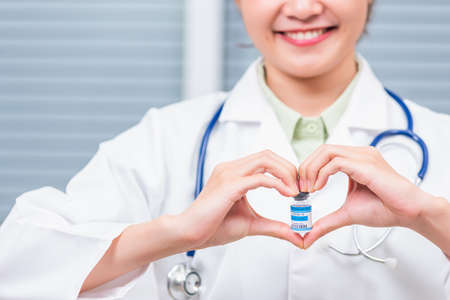Closeup woman doctor smile forming a heart with her hands mark heart finger showing a glass vial of coronavirus vaccine,  COVID-19 Medicine And Health Care Conceptの写真素材