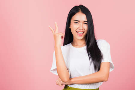 Asian happy portrait beautiful cute young woman teen smile standing show finger making v-sign victory sign gesture side away looking to camera studio shot isolated on pink background with copy spaceの写真素材