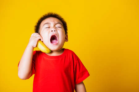 Happy portrait Asian child or kid cute little boy attractive smile wearing red t-shirt playing holds banana fruit pretending to be like a telephone, studio shot isolated on yellow backgroundの写真素材