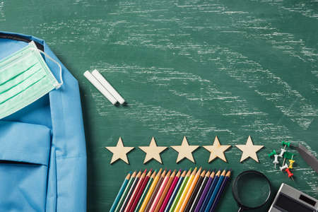 Top view of stylish blue school bag backpack at a green chalkboard with face mask protection and...