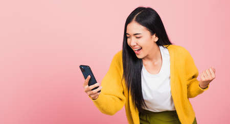 Happy Asian portrait beautiful cute young woman teen smiling excited   using mobile phone say yes! Studio shot isolated on pink background, Thai female surprised make winner gesture on smartphoneの写真素材