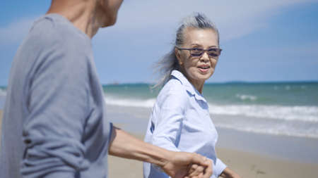 Happy Asian senior man and woman couple holding hands walking to the beach sunny with bright blue sky, Romantic elderly enjoy Travel summer vacation, plan life insurance at retirement couple conceptの写真素材