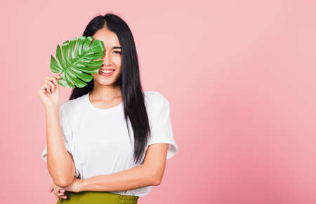 Beauty face. Portrait of Asian beautiful young woman with fresh healthy skin hold green monstera leaf on her face, Tropical Leaves, studio shot isolated on pink background, Skin body care spa conceptの写真素材