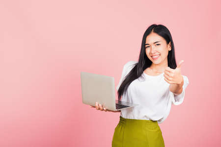 Portrait of happy Asian beautiful young woman confident smiling face holding using laptop computer and showing thumb up for like gesture, studio shot with copy space isolated on pink backgroundの写真素材