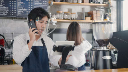Happy Asian barista man coffee maker receive phone call and record pre order to computer in coffee cafe shop, Manager service small business conceptの写真素材