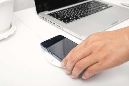 Woman putting smartphone charging power battery with wireless charger device near laptop computer,...