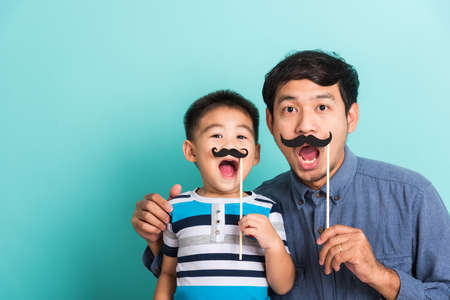 Family funny happy hipster father and his son kid holding black mustache props for the photo booth close face, studio shot isolated on a blue background, November men health awarenessの写真素材