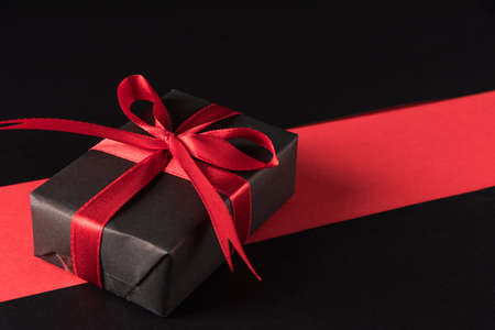 Black Friday sale shopping and Boxing Day concept, Gift box wrapped black paper and red bow ribbon present, studio shot isolated on red and dark backgroundの写真素材