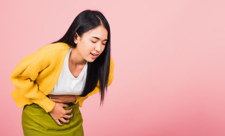 Portrait of Asian beautiful young woman has stomachache, female abdominal pain suffering from stomach ache, studio shot isolated on pink background, Health and medical gastritis conceptの写真素材