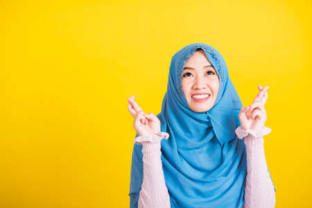 Asian Muslim Arab, Portrait of happy Asian beautiful young woman Islamic religious wear veil hijab she smiling and holding fingers crossed for good luck, studio shot isolated on yellow backgroundの写真素材