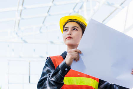Asian engineer architect worker woman holding blueprint infrastructure progress at construction site, engineering female leader standing hold paper plan control worker to building projectの写真素材