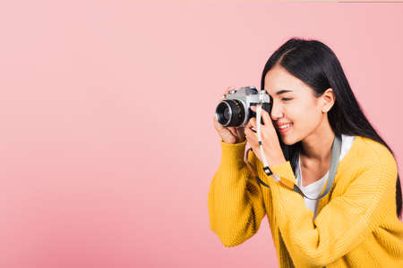 Attractive energetic happy Asian portrait beautiful young woman smiling photographer taking a picture and looking viewfinder on retro vintage photo camera ready to shoot isolated on pink backgroundの写真素材