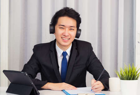 Work from home, Asian young businessman wear suit video conference call or facetime he smiling sitting on desk using smart digital tablet computer and write paper notebook information at home officeの写真素材