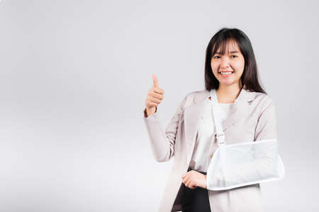 Arm broken. Business woman confident smile broken after accident wear arm splint for treatment show thumb up finger for good sign, happy Asian female sling support hand isolated on white backgroundの写真素材