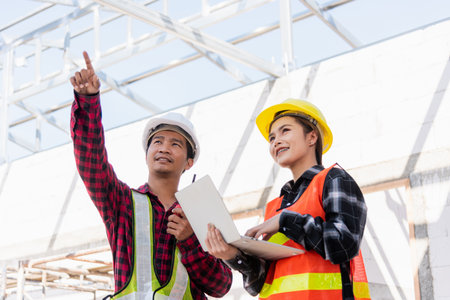 Asian engineer foreman worker man and woman working at building construction site use laptop and talking with radio, engineering hold computer and radio discuss and control worker employee to buildingの写真素材