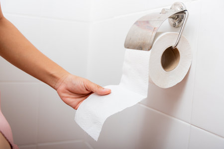 Closeup hand pulling toilet paper roll in holder for wipe, woman sitting on toilet she taking and tearing white tissue on wall to towel clean in bathroom, Healthcare conceptの写真素材
