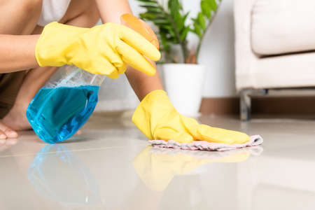 Asian woman wearing yellow rubber glover with cloth rag and detergent spray cleaning floor at home in living room, Female hands wash cleaner, housework and housekeeping conceptの写真素材