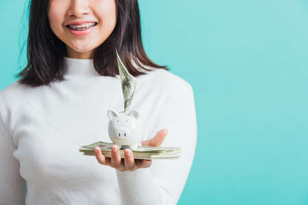 Asian beautiful young woman holding piggy bank on hands and putting dollars money banknotes, portrait of happy female smiling hold piggybank in palm, isolated on blue background, saving money conceptの写真素材