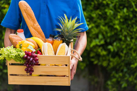 Asian man farmer wears delivery uniform he holding full fresh vegetables and fruits in crate wood box in hands ready give to customer harvest organic food on the garden place green leaves backgroundの写真素材