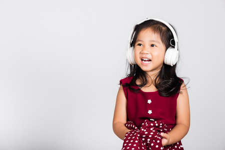Little cute kid girl 3-4 years old listen music in wireless headphones in studio shot isolated on white background, happy Asian children smiling listening audio, entertainment lifestyleの写真素材