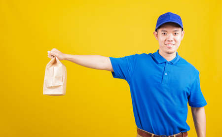 Portrait excited delivery service man smile wearing blue t-shirt and cap uniform hold paper containers for takeaway bag grocery food packet looking to camera, studio shot isolated on yellow backgroundの写真素材