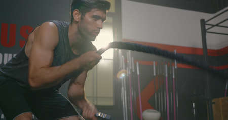 Muscular athletic Caucasian young man strong effort with battle rope waves doing exercise while...