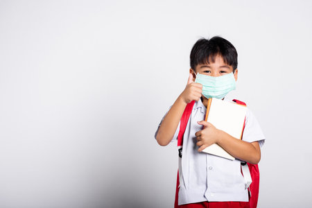 Asian student kid boy wearing student thai uniform and medical protect face mask and show thumb up finger for good in studio shot isolated on white background, new normal back to schoolの写真素材