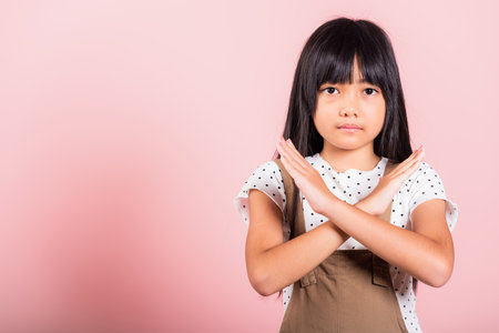 Stop gesture. Asian little kid 10 years old showing two hands cross arms say no X sign at studio shot isolated on pink background, Happy child girl disagreement oppositionの写真素材