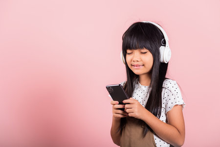 Asian kid 10 years enjoying listening music from mobile phone wear wireless headset closed eyes at studio shot isolated on pink background, Happy child girl use smartphone listen music with headphonesの写真素材