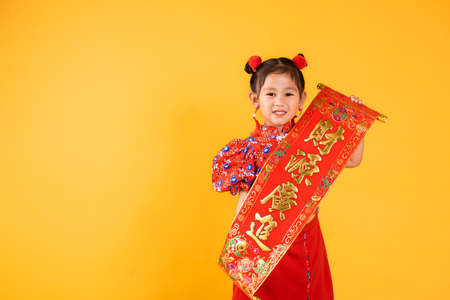 Chinese New Year. Happy Asian Chinese little child girl smile wearing red cheongsam qipao Spring festival couplets (Character "FU" means fortune, blessing), studio short isolated on yellow backgroundの写真素材