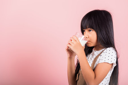 Asian little kid 10 years old smile hold milk glass drink white milk and drinking at studio shot isolated on pink background, Happy child girl daily life health care Medicine foodの写真素材