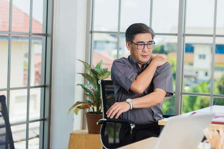 Asian hard senior businessman working with laptop computer has a problem with shoulder pain. Old man feeling pain after sitting at desk long time, Healthcare and medicine office syndrome conceptの写真素材