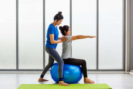 Two Asian adult female and trainer young woman in sportswear doing aerobics yoga exercise with sitting on fitness ball indoor yoga studio, fit sport healthy workout lifestyle exerciseの写真素材