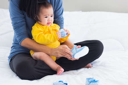 Asian young mother sitting on the bed wearing shoes to his cute baby little girl toddler on bedroom, Mom helping little infant wears shoesの写真素材