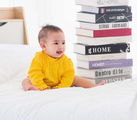Asian cute baby little girl toddler looking at a stack of books on bedroom white bed, First baby library conceptの写真素材