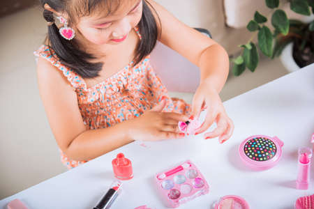 Asian adorable funny little girl making makeup dips brush into bottle to paints nails polish red nail varnish herself, Learning activity to be woman, happy kid is beautiful make up with cosmetics toyの写真素材