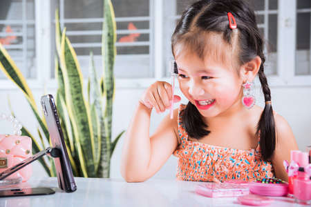 Asian funny little girl making makeup her face she look in smartphone and use eyebrow mascara, Learning activity to be woman, social media blogger recording vlog with cosmetics, online tutorialの写真素材