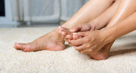 Foot pain, Asian woman holds her toe injury feeling pain her foot at home, female suffering from feet ache use hand massage relax muscle from toe in house interior, Healthcare problems medical conceptの写真素材