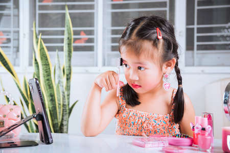 Asian funny little girl making makeup her face she look in smartphone and use eyebrow mascara, Learning activity to be woman, social media blogger recording vlog with cosmetics, online tutorialの写真素材