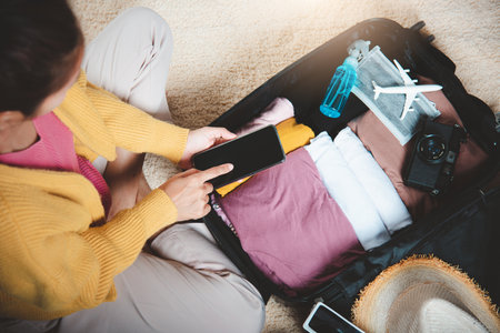 Woman using smartphone blank screen and packing suitcase bag for travel trip weekend vacation at home, female check information in tourism website on mobile phone before go to airportの写真素材