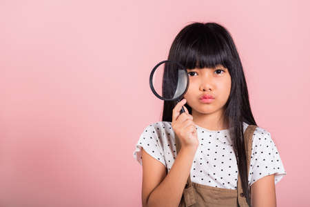 Asian little kid 10 years old funny looking through magnifying glass at studio shot isolated on pink background, Happy child girl lifestyle smiling exploring holding magnifier searchingの写真素材