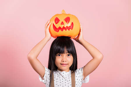 Asian little kid 10 years old holding carved Halloween pumpkin at studio shot isolated on pink background, Portrait of happy child girl smiling show Jack O pumpkin, Concept for Halloween holidayの写真素材