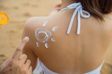 Happy young man applying suntan lotion with sun shape on girlfriend back shoulder at the beach,...