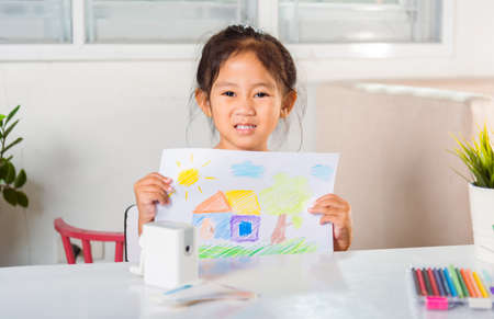 Happy child little girl lift up colorful drawing landscape my home dream on paper to camera, Asian cute kid preschooler smiling showing draw country house picture at home to learning arts homeworkの写真素材